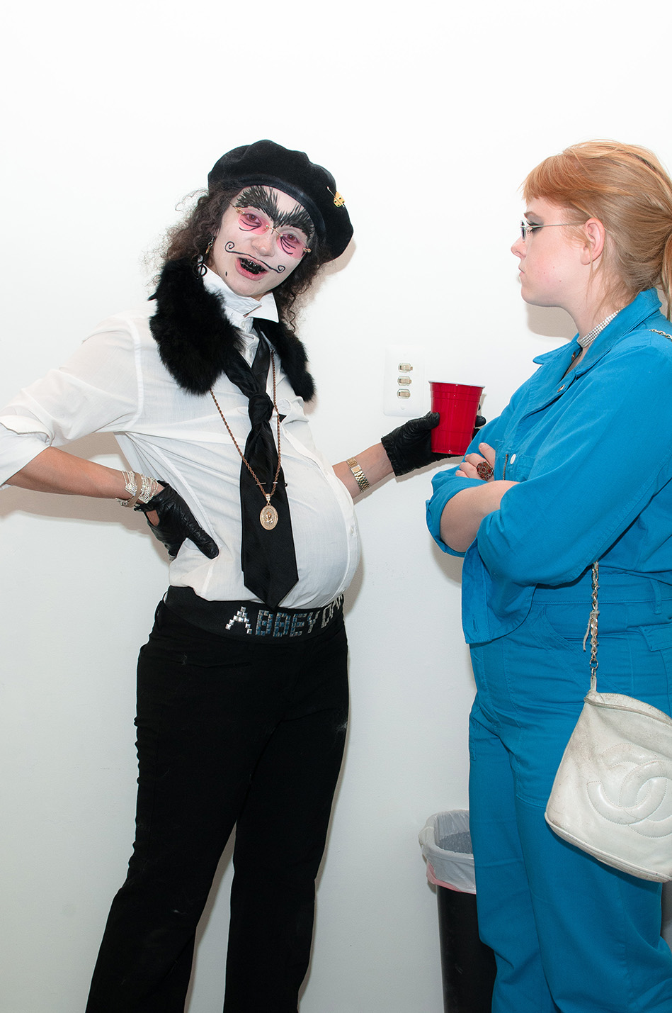A pregnant figure with a drawn on mustache and unibrow and blacked out teeth faces the camera